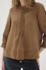 Blusa de lino color marrón, de corte recto y mangas 3/4. Presenta escote redondo con pequeños volados y pliegues frontales, además de detalles de encaje en las mangas y puños con volados.