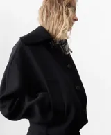 Chaqueta negra de corte corto y holgado, confeccionada con mezcla de lana. Cuenta con cuello ancho y solapa de piel sintética en tono beige claro, cierre frontal con botones oscuros y mangas largas amplias.