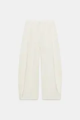 Pantalón fluido de color blanco, de tiro alto con trabillas y pliegues en el delantero. Cuenta con bolsillos laterales, cierre frontal con cremallera, botón interior y gancho metálico.