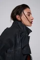 Campera negra oversized estilo utilitario con botones metálicos, hombros caídos, bolsillos con solapa y terminación levemente fruncida en el ruedo. El acabado tipo cuero aporta textura y un carácter urbano distintivo.