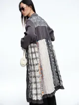 Chaqueta larga de diseño patchwork en tonos grises, con combinación de texturas que incluyen paneles de tela lisa, tela texturizada blanca y paneles con estampado floral en relieve. Presenta cierre frontal con botones, cuello camisero y bordes deshilachados.