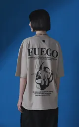 Remera de manga corta y cuello redondo con calce oversize, color beige con estampa gráfica en negro que incluye texto y un rostro femenino estilizado con llamas.