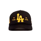 Gorro trucker negro con diseño de red en la parte trasera, visera curva y bordado frontal en hilo amarillo con las letras 'LA' y las leyendas 'Mi CASA' y 'Su CASA'.