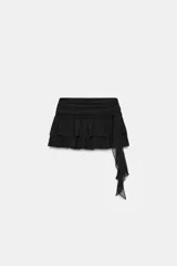 Falda pantalón de tiro medio color negro, con diseño de volantes y tejido asimétrico lateral que incluye un lazo colgante. Cuenta con cierre lateral de cremallera oculta.