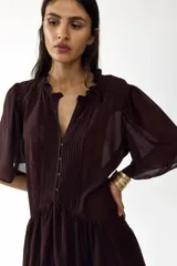 Vestido corto de color bordó, con escote en V y cuello de volados, mangas cortas abullonadas y botonadura frontal con alforzas e inserciones de encaje. Presenta costuras angulares con frunces en la cadera y falda con ribete de volado en el bajo.