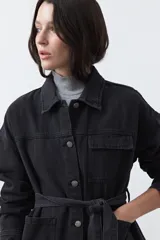 Sobrecamisa de jean color negro con silueta relajada, cuello camisero, cierre frontal con botones metálicos, bolsillos frontales y cinturón ajustable en la cintura.