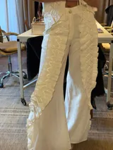 Pantalón de lino color blanco, de corte amplio y tiro medio. Presenta detalles de tablas verticales a lo largo de las piernas con costuras decorativas en zigzag, bolsillos laterales y traseros, y aberturas laterales.