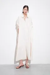 Vestido camisero largo de manga larga, color blanco con finas rayas verticales en tono claro, con cuello solapa y abotonadura frontal. Posee un calce amplio y bolsillos laterales.