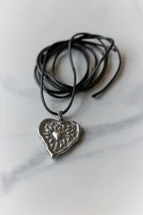 Collar con dije de corazón en acero quirúrgico, con textura irregular y relieve, montado sobre un tiento encerado negro.