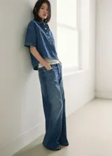 Blusa de algodón estilo denim, con diseño oversize, cuello camisero, mangas cortas, dos bolsillos de parche en el pecho y cierre frontal de botones.