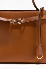 Cartera de hombro color tostado, fabricada en tejido revestido, con correa ajustable y un divisor interior que crea dos compartimentos, además de un compartimento interior con cierre. Presenta una trabilla decorativa con mosquetón metálico dorado en la parte delantera y una etiqueta extraíble.