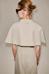Vestido camisero midi de algodón con diseño de capa, cuello solapa, manga corta tipo capa y trabillas en los hombros. Presenta cierre frontal con botones, bolsillos delanteros y cinturón ajustable del mismo tejido con lazada. El bajo tiene un acabado en línea evasé.
