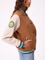 Campera estilo varsity con cuerpo en color marrón y mangas en tono beige. Presenta cierre frontal con botones a presión, cuello, puños y ruedo con franjas tejidas en verde y blanco. Incluye un parche con la letra R en el pecho y detalles bordados en las mangas.