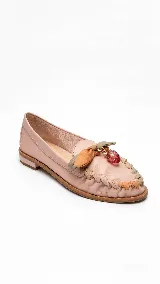 Mocasín de cuero color nude con punta redondeada y taco bajo. Presenta un detalle artesanal de tiras entrelazadas en tonos verde, marrón y beige en el empeine, acompañado por un moño y un dije colgante en forma de frutilla de cristal.