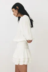 Vestido corto de color blanco en tejido con textura, con escote redondo, abertura con tiras para anudar, mangas largas abullonadas y falda de volados escalonados.