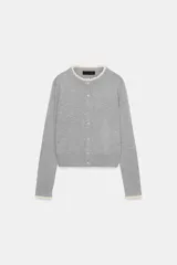 Chaqueta de punto de corte clásico, manga larga y cuello redondo. Presenta cierre frontal con botones y detalles de puntilla en contraste en el cuello y los puños.
