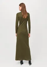 Vestido largo de punto en color verde oliva, con cuello alto y manga larga. Presenta un diseño ajustado en la parte superior que se extiende hacia una falda recta, con un detalle metálico decorativo en el lateral de la cintura.
