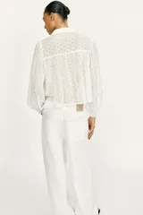 Blusa corta de color blanco con diseño de bordados perforados, cuello solapa, cierre frontal con botones y mangas largas abullonadas.