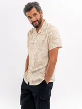 Camisa de manga corta color beige con estampado tropical de hojas, cuello camisero abierto y cierre frontal de botones.