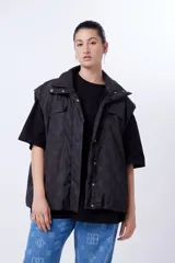 Chaleco negro con estampado de logos, cuello alto, cierre frontal con cremallera y broches a presión ocultos, bolsillos delanteros y cintura ajustable con cordón elástico. Interior de tafeta relleno de guata.