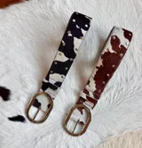 Cinturón de cuero vacuno con pelo, estampado de manchas tipo vaca, hebilla ovalada plateada y detalles de tachas metálicas en los bordes.