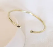 Pulsera rígida abierta en color dorado, con iniciales grabadas en los extremos.