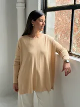 Sweater de punto color beige, de corte holgado y diseño asimétrico con tajos laterales. Presenta cuello redondo y mangas largas con puños acanalados, además de un detalle de costura vertical central.