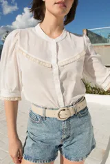 Camisa de estilo romántico color blanco, confeccionada en tejido liviano con textura de rayas verticales. Presenta mangas al codo con frunces en los hombros, cuello redondo y cierre frontal con botones. Incluye delicados detalles de puntilla de algodón en el pecho y en los bordes de las mangas.
