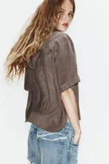 Camisa de corte fluido con cuello solapa y manga corta con dobladillo. Presenta un cierre frontal con botones ocultos bajo una tapeta y un diseño con bajo ligeramente más largo en la parte posterior.