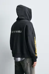 Sudadera tipo hoodie de corte relajado, color negro, con capucha y bolsillo frontal tipo canguro. Presenta parches decorativos en el pecho y estampados en color amarillo en la manga derecha. Acabados en rib en puños y ruedo.
