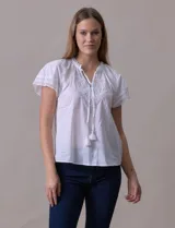 Blusa blanca de algodón marca Cupio, con cuello a la base, escote en V y lazo de ajuste. Presenta bordado en la parte delantera y mangas cortas amplias con aplique de puntilla.