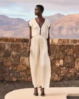 Vestido largo color crudo, confeccionado en viscosa satinada de inspiración grecorromana. Cuenta con escote en V profundo con ribete negro en contraste, mangas cortas fluidas con detalles oscuros, y corte en la cintura con drapeado suave.