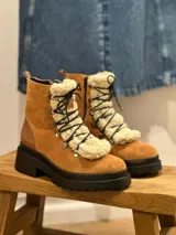 Bota tipo borcego de cuero color suela, con suela track negra de goma. Presenta cordones negros y un aplique de corderito sintético desmontable en la lengüeta.