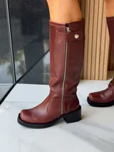 Bota de caña alta estilo biker, confeccionada en cuero color chocolate. Presenta un cierre metálico lateral que recorre toda la caña, estribo y cadena decorativa desmontables, y taco cuadrado con plataforma.