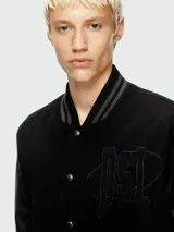 Campera estilo universitaria (varsity jacket) negra con mangas de cuero sintético negro y cuerpo de lana negra. Presenta un parche bordado grande en el pecho y cierre frontal de botones a presión. Tiene cuello, puños y cintura acanalados.