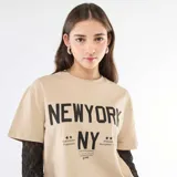 Camiseta de manga corta color beige con estampado gráfico frontal en negro, que incluye una segunda capa de mangas largas de encaje negro debajo.