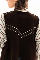 Chaleco oversize confeccionado en cuero descarne, con diseño de terminación en picos y cierre frontal con lazos y ojalillos. Presenta tachas metálicas decorativas en los hombros y en la parte posterior.