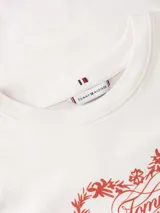 Remera de algodón de corte regular, color blanco, con cuello redondo y mangas cortas. Presenta un estampado frontal en color rojo que combina un diseño floral en forma de corazón con el logo de la marca en el centro. Incluye el logo de la marca bordado en la manga.