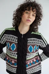 Campera de punto jacquard con lana merino, diseño con cierre frontal, cuello redondo y detalles de trenzas en los laterales. Presenta un patrón geométrico multicolor con tonos marrones, negros, azules y verdes.