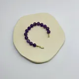 Pulsera compuesta por cuentas esféricas de piedra amatista natural de 10 mm, unidas con nudos separadores en hilo color violeta. Presenta cierre de mosquetón y detalles metálicos con baño de oro.