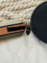 Monedero circular de color negro con cierre perimetral. Presenta una etiqueta frontal con el logo de la marca y una correa lateral de tela con franjas en tonos beige, marrón y negro.