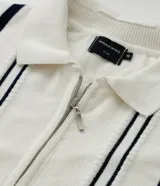 Camisa polo slim de punto, color blanco con rayas verticales oscuras en contraste en el frente y líneas en los bordes de las mangas cortas. Cuenta con cuello polo y cierre frontal parcial con cremallera.