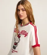Remera blanca de algodón con cuello redondo y mangas cortas con ribetes rojos. Estampado frontal con el logo de Hello Kitty y detalles de rayas rojas en las mangas.