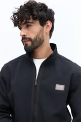 Campera negra de algodón con cierre frontal completo, cuello alto y bolsillos laterales con botón a presión. Presenta un pequeño parche rectangular con texto en el pecho izquierdo.
