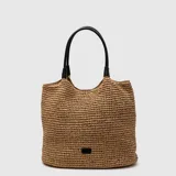 Bolso tipo tote confeccionado en rafia tejida, con manijas largas de cuero sintético en color negro. Presenta una estructura amplia y un pequeño aplique metálico con logo en la parte inferior frontal.