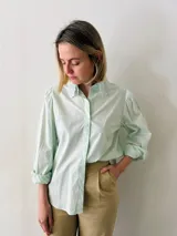 Camisa verde agua con cuello babero de puntilla blanca, mangas largas abullonadas con puños elastizados y cierre frontal con botones.