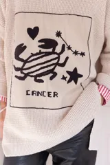 Sweater de punto oversize color crudo, con cuello redondo y diseño bordado a mano en el frente representando al signo Cáncer con un cangrejo, un corazón y una estrella.
