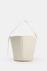 Bolso de hombro de piel en color blanco, con diseño tipo bucket, asa fija y detalle de tira colgante con nudo en el lateral.