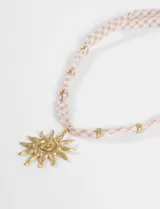 Collar con dije de sol en tono dorado y cordón de tela con estampado de cuadros vichy en tonos rosados. Presenta pequeñas cuentas metálicas doradas distribuidas a lo largo del cordón.