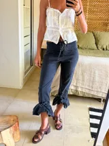 Pantalón de jean oscuro, tiro alto, con cordón de ajuste frontal a modo de corsé. El ruedo es capri y termina en un gran volado fruncido y anudado.